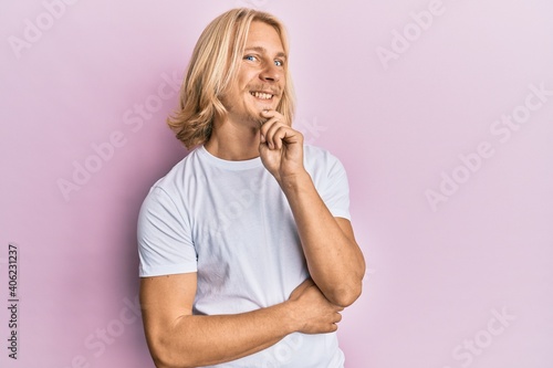 Caucasian young man with long hair wearing casual white t shirt looking confident at the camera with smile with crossed arms and hand raised on chin. thinking positive.