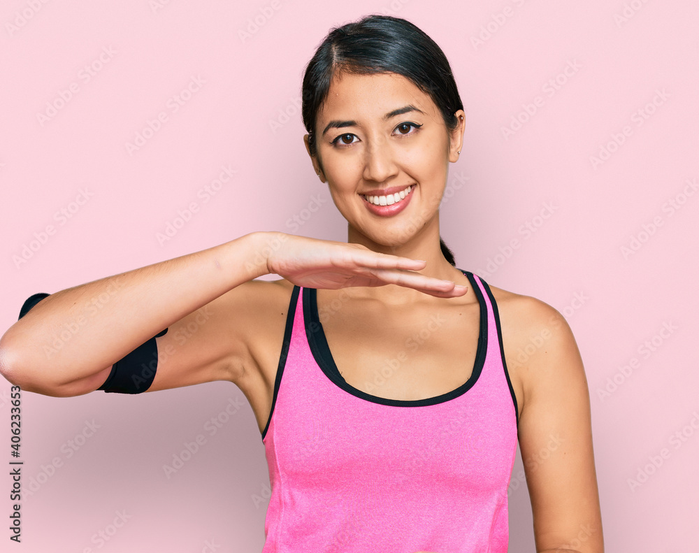 Beautiful asian young sport woman wearing sportswear and arm band gesturing with hands showing big and large size sign, measure symbol. smiling looking at the camera. measuring concept.