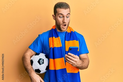 Young caucasian man holding football ball looking at smartphone afraid and shocked with surprise and amazed expression  fear and excited face.