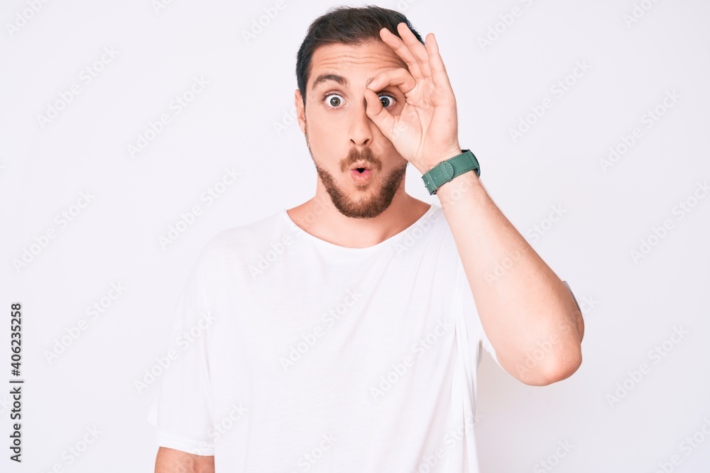 Young handsome man wearing casual white tshirt doing ok gesture shocked with surprised face, eye looking through fingers. unbelieving expression. Young handsome man wearing casual white tshirt doing ok gesture shocked with surprised face, eye looking through fingers. unbelieving expression.