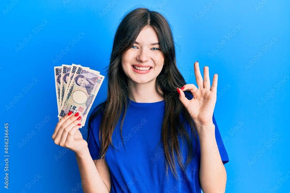 Young beautiful caucasian girl holding 5000 japanese yen banknotes doing ok sign with fingers, smiling friendly gesturing excellent symbol Young beautiful caucasian girl holding 5000 japanese yen banknotes doing ok sign with fingers, smiling friendly gesturing excellent symbol
