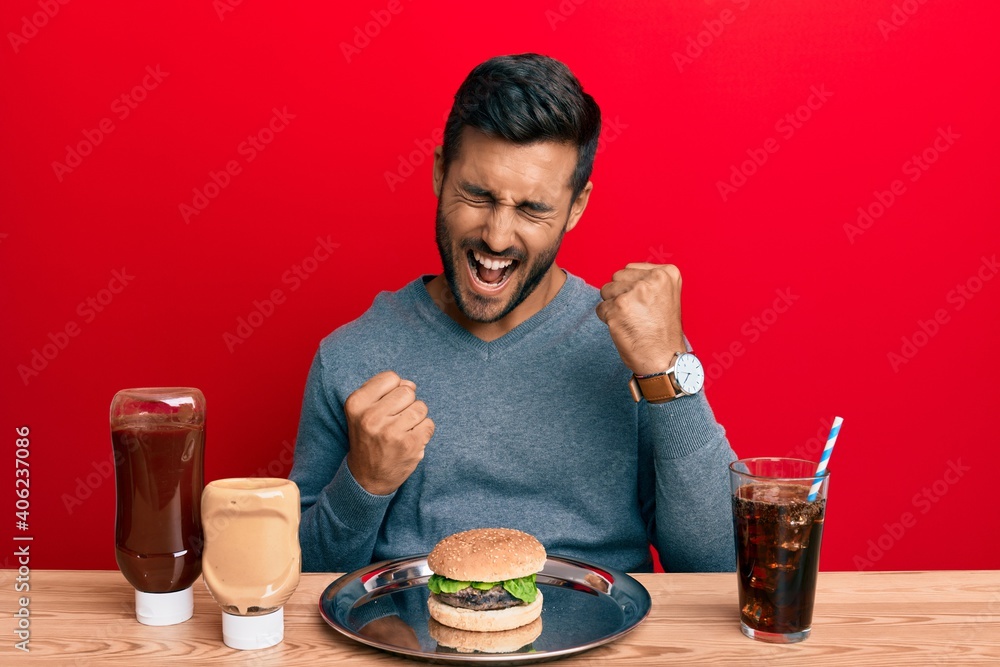 Handsome hispanic man eating a tasty classic burger and soda celebrating surprised and amazed for success with arms raised and eyes closed Handsome hispanic man eating a tasty classic burger and soda celebrating surprised and amazed for success with arms raised and eyes closed