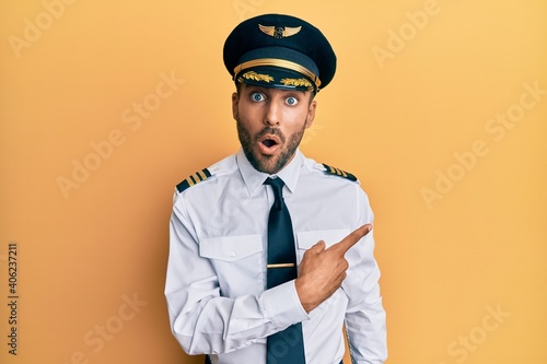 Handsome hispanic man wearing airplane pilot uniform surprised pointing with finger to the side, open mouth amazed expression Fototapet