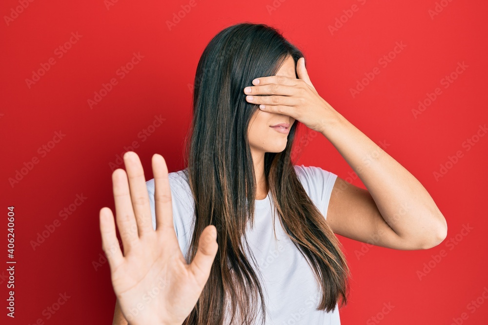 Young brunette woman wearing casual white tshirt over red background covering eyes with hands and doing stop gesture with sad and fear expression. embarrassed and negative concept. Young brunette woman wearing casual white tshirt over red background covering eyes with hands and doing stop gesture with sad and fear expression. embarrassed and negative concept.