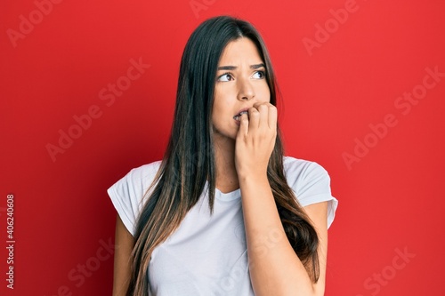 Young brunette woman wearing casual white tshirt over red background looking stressed and nervous with hands on mouth biting nails. anxiety problem.