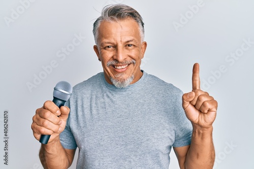 Middle age grey-haired man singing song using microphone smiling with an idea or question pointing finger with happy face, number one