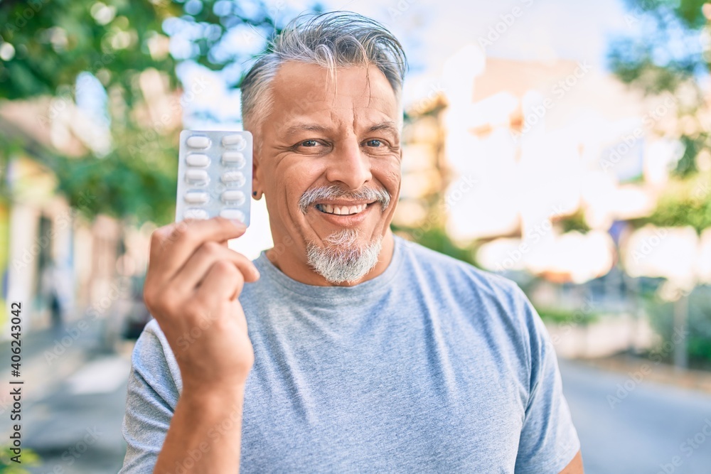 Middle age hispanic grey-haired man smiling happy holding pills standing at the city. Middle age hispanic grey-haired man smiling happy holding pills standing at the city.