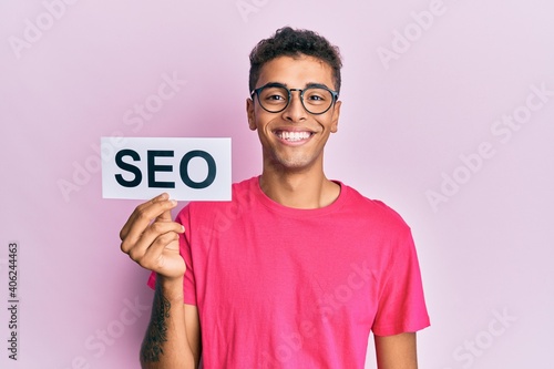 Young handsome african american man holding seo message paper looking positive and happy standing and smiling with a confident smile showing teeth