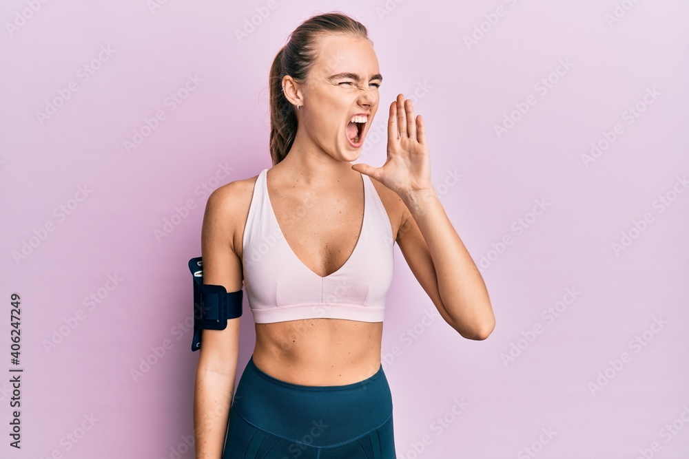 Beautiful blonde woman wearing sportswear and arm band shouting and screaming loud to side with hand on mouth. communication concept.
