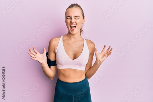 Beautiful blonde woman wearing sportswear and arm band crazy and mad shouting and yelling with aggressive expression and arms raised. frustration concept. © Krakenimages.com