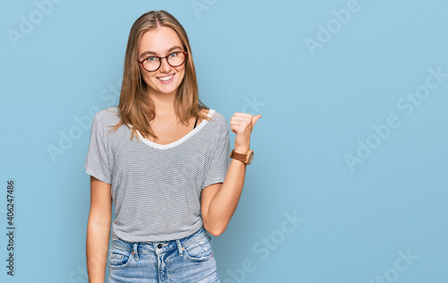 Beautiful young blonde woman wearing casual clothes and glasses smiling with happy face looking and pointing to the side with thumb up.