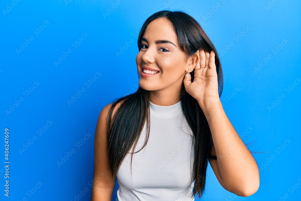 Young beautiful hispanic girl wearing casual clothes smiling with hand over ear listening and hearing to rumor or gossip. deafness concept. Young beautiful hispanic girl wearing casual clothes smiling with hand over ear listening and hearing to rumor or gossip. deafness concept.