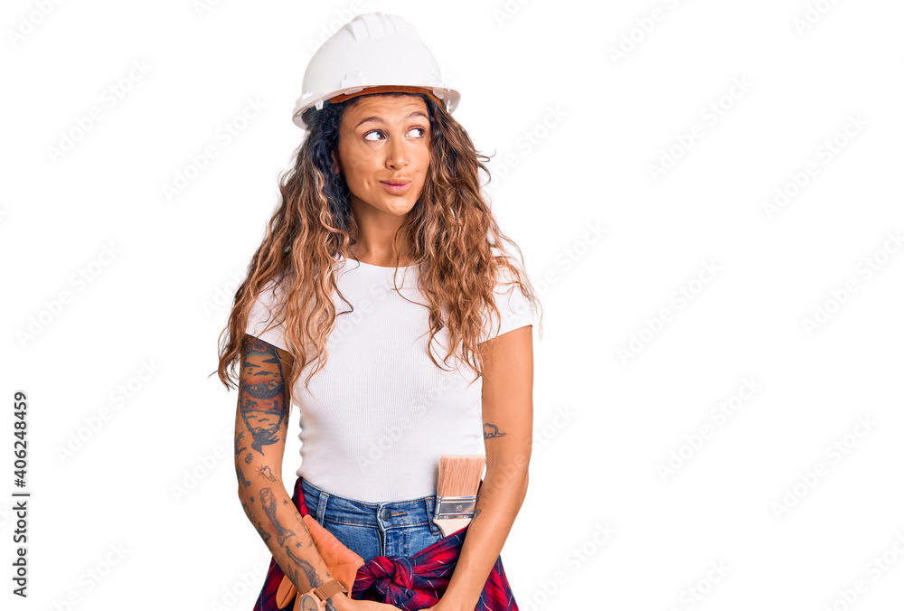 Young hispanic woman with tattoo wearing hardhat and builder clothes smiling looking to the side and staring away thinking. Young hispanic woman with tattoo wearing hardhat and builder clothes smiling looking to the side and staring away thinking.