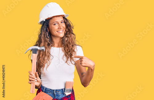 Young hispanic woman with tattoo wearing hardhat and builder clothes holding hammer pointing finger to one self smiling happy and proud