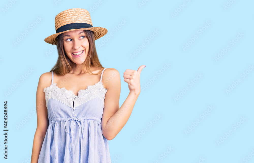 Beautiful brunette young woman wearing summer hat smiling with happy face looking and pointing to the side with thumb up.