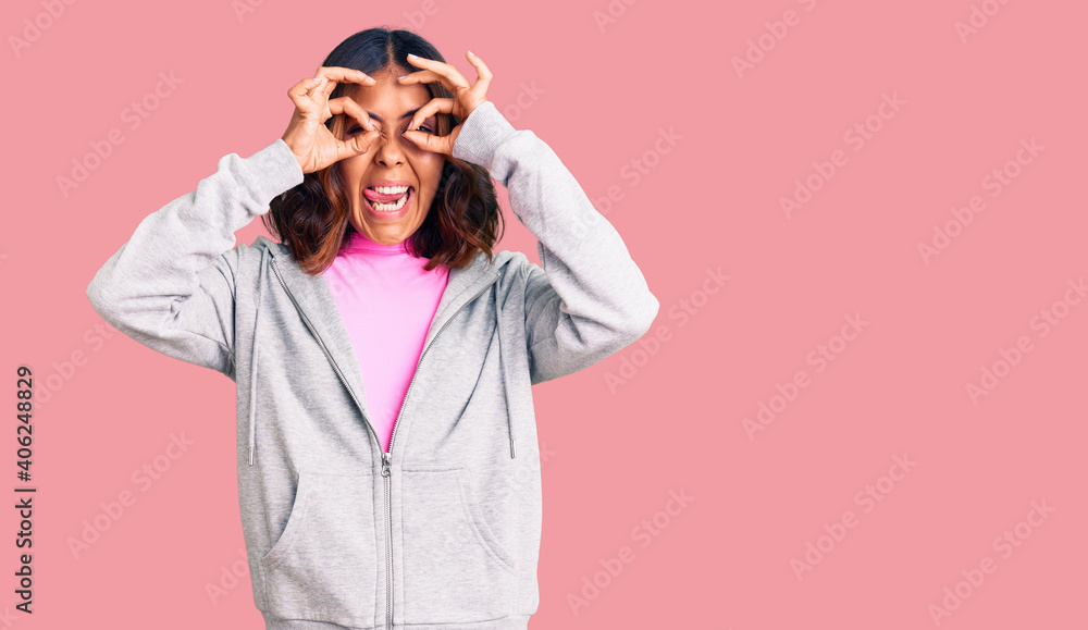Young beautiful mixed race woman wearing casual sporty clothes doing ok gesture like binoculars sticking tongue out, eyes looking through fingers. crazy expression. Young beautiful mixed race woman wearing casual sporty clothes doing ok gesture like binoculars sticking tongue out, eyes looking through fingers. crazy expression.