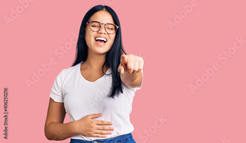 Young beautiful asian girl wearing casual clothes and glasses laughing at you, pointing finger to the camera with hand over body, shame expression