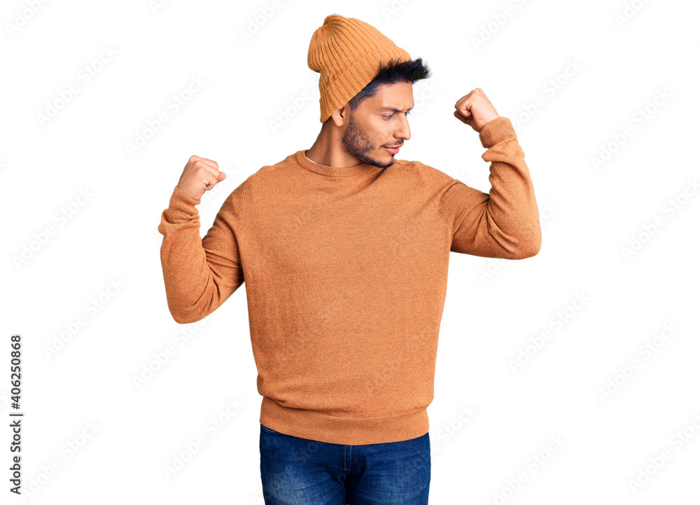 Naklejka premium Handsome latin american young man wearing winter sweater and wool hat showing arms muscles smiling proud. fitness concept.