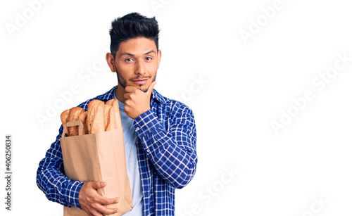 Handsome latin american young man holding paper bag with bread looking confident at the camera smiling with crossed arms and hand raised on chin. thinking positive.