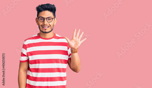 Handsome latin american young man wearing casual clothes and glasses showing and pointing up with fingers number five while smiling confident and happy.
