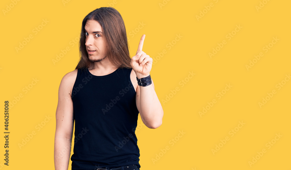 Young adult man with long hair wearing goth style with black clothes pointing finger up with successful idea. exited and happy. number one.