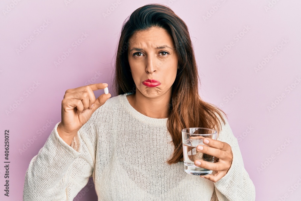 Beautiful hispanic woman holding pill and glass of water depressed and worry for distress, crying angry and afraid. sad expression. Beautiful hispanic woman holding pill and glass of water depressed and worry for distress, crying angry and afraid. sad expression.