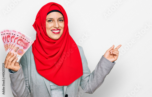 Beautiful hispanic woman wearing islamic hijab holding chinese yuan banknotes smiling happy pointing with hand and finger to the side