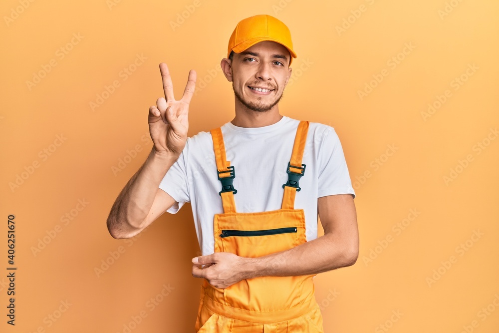 Hispanic young man wearing handyman uniform smiling looking to the camera showing fingers doing victory sign. number two. Hispanic young man wearing handyman uniform smiling looking to the camera showing fingers doing victory sign. number two.