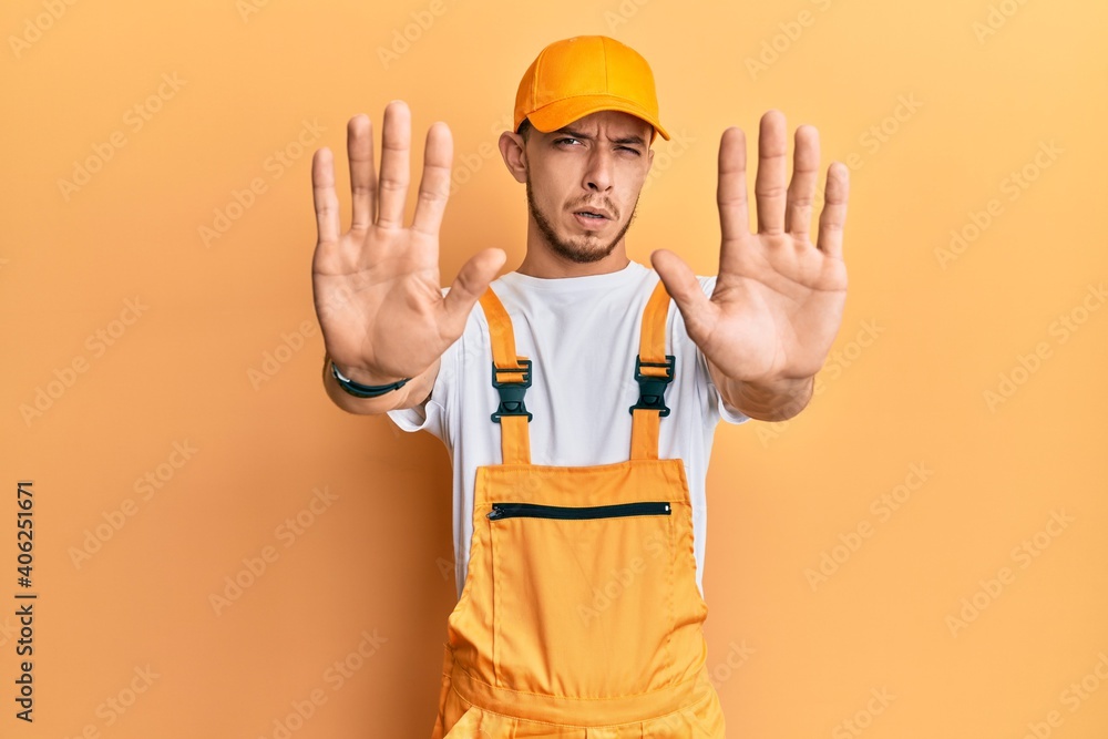 Hispanic young man wearing handyman uniform doing stop gesture with hands palms, angry and frustration expression