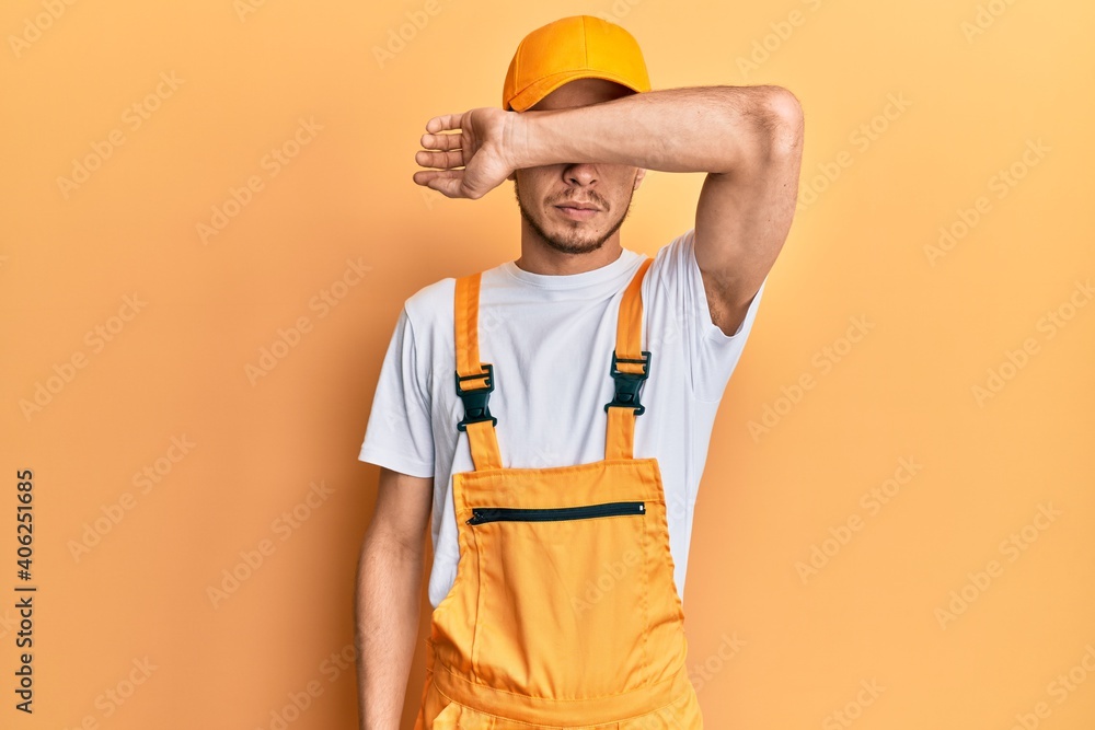 Hispanic young man wearing handyman uniform covering eyes with arm, looking serious and sad. sightless, hiding and rejection concept