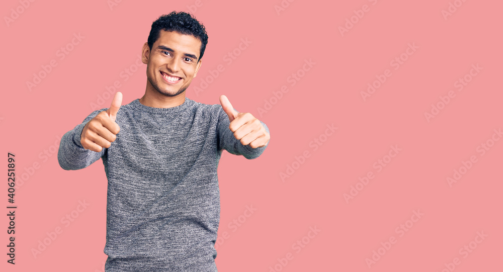 Hispanic handsome young man wearing casual sweater approving doing positive gesture with hand, thumbs up smiling and happy for success. winner gesture. Hispanic handsome young man wearing casual sweater approving doing positive gesture with hand, thumbs up smiling and happy for success. winner gesture.