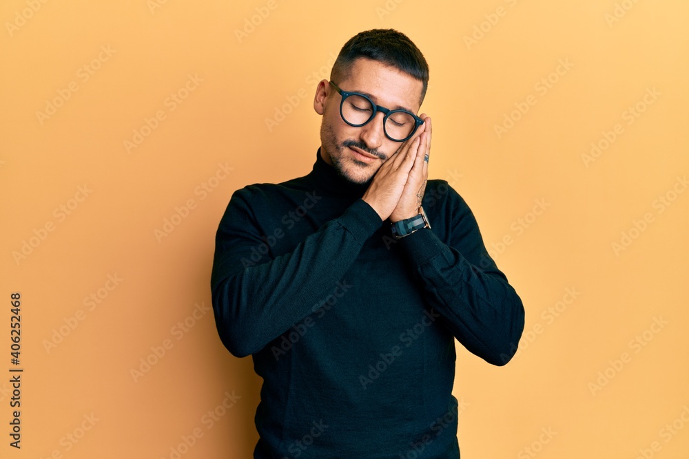 Handsome man with tattoos wearing turtleneck sweater and glasses sleeping tired dreaming and posing with hands together while smiling with closed eyes. Handsome man with tattoos wearing turtleneck sweater and glasses sleeping tired dreaming and posing with hands together while smiling with closed eyes.