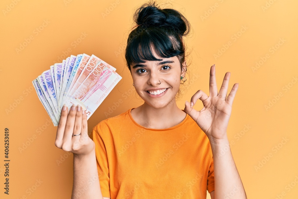 Young hispanic girl holding swedish krona banknotes doing ok sign with fingers, smiling friendly gesturing excellent symbol Young hispanic girl holding swedish krona banknotes doing ok sign with fingers, smiling friendly gesturing excellent symbol