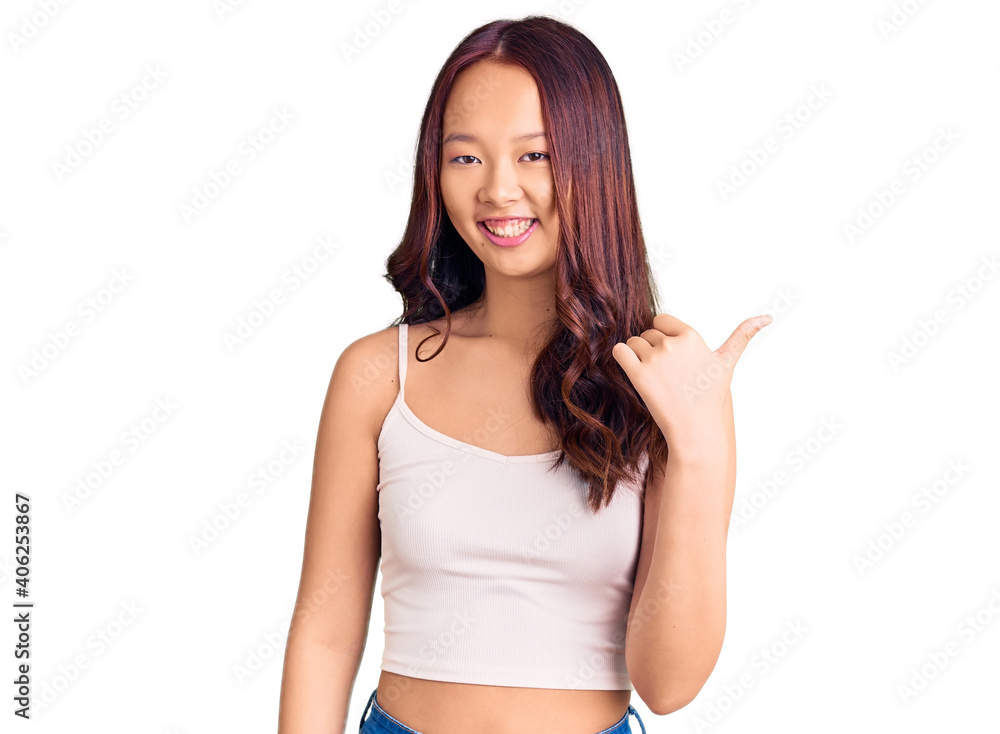 Young beautiful chinese girl wearing casual clothes smiling with happy face looking and pointing to the side with thumb up. Young beautiful chinese girl wearing casual clothes smiling with happy face looking and pointing to the side with thumb up.