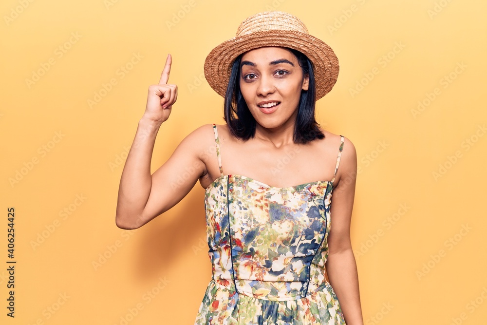 Young beautiful latin woman wearing summer hat pointing finger up with successful idea. exited and happy. number one. Young beautiful latin woman wearing summer hat pointing finger up with successful idea. exited and happy. number one.