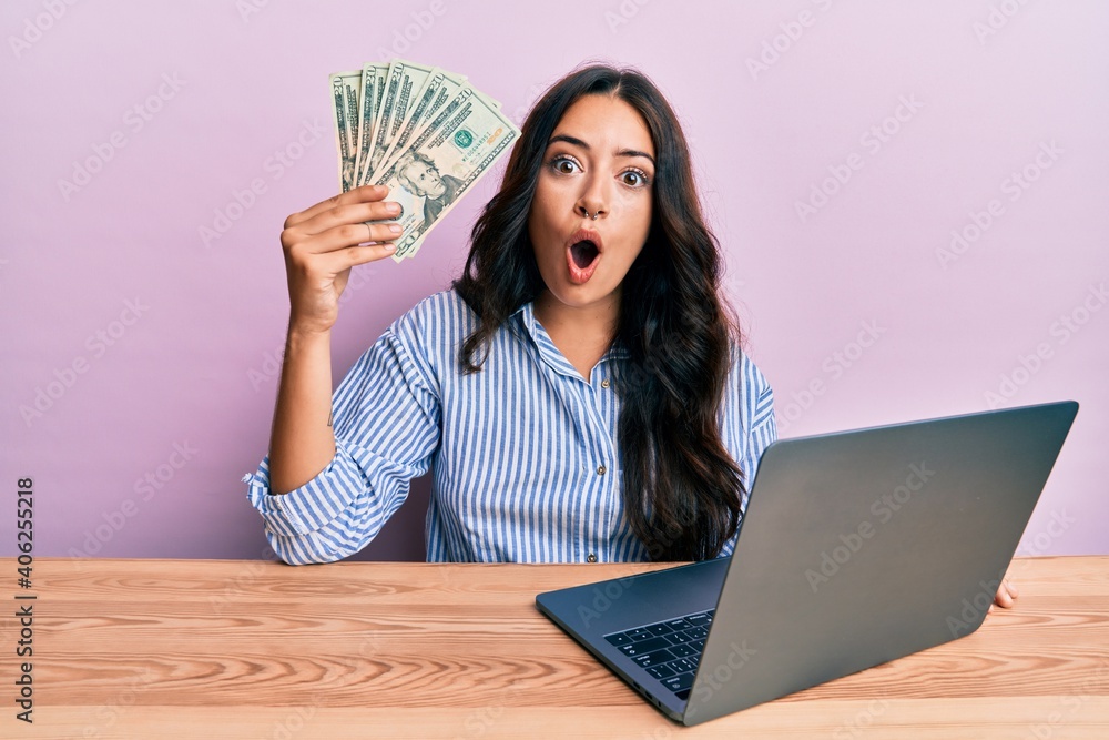 Beautiful brunette young woman working at the office holding dollars scared and amazed with open mouth for surprise, disbelief face Beautiful brunette young woman working at the office holding dollars scared and amazed with open mouth for surprise, disbelief face