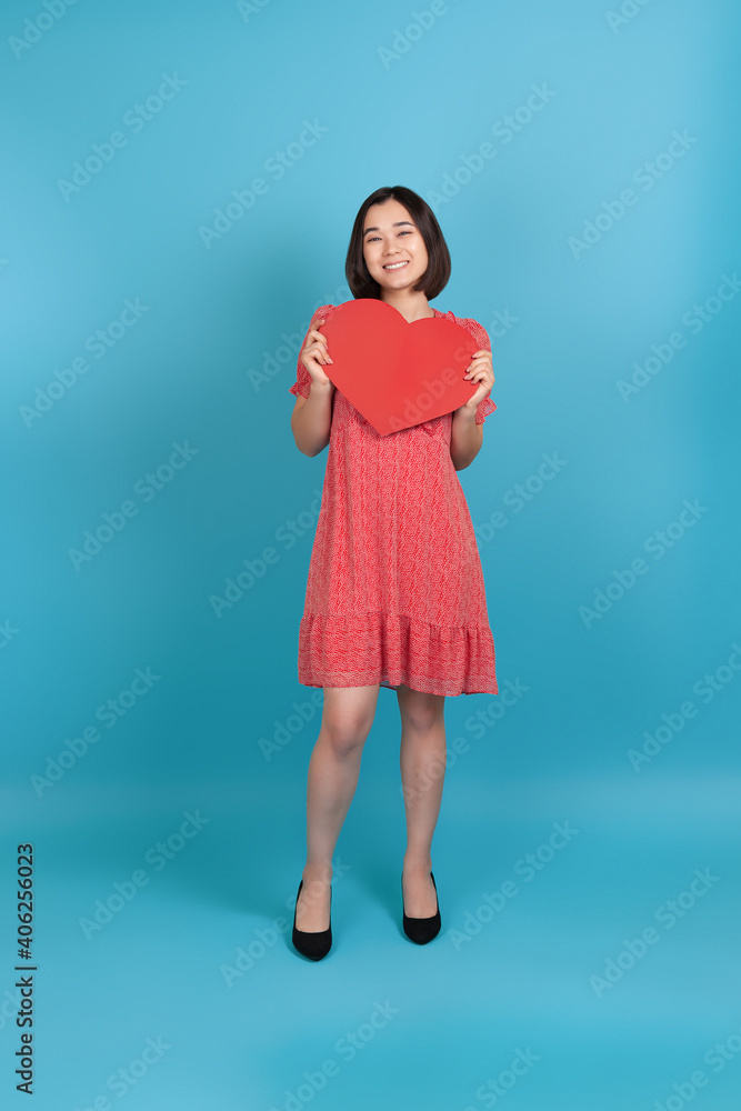 full-length smiling happy young asian woman in red dress holding big red paper heart isolated on blue background. full-length smiling happy young asian woman in red dress holding big red paper heart isolated on blue background.