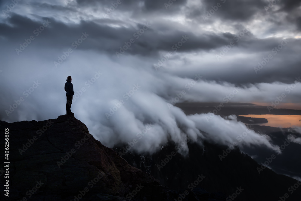 Man Hiker on top of a Mountain Peak. Dreamscape Artistic Render Composite. Landscape background from British Columbia, Canada. Dark Dramatic Sunset Sky.