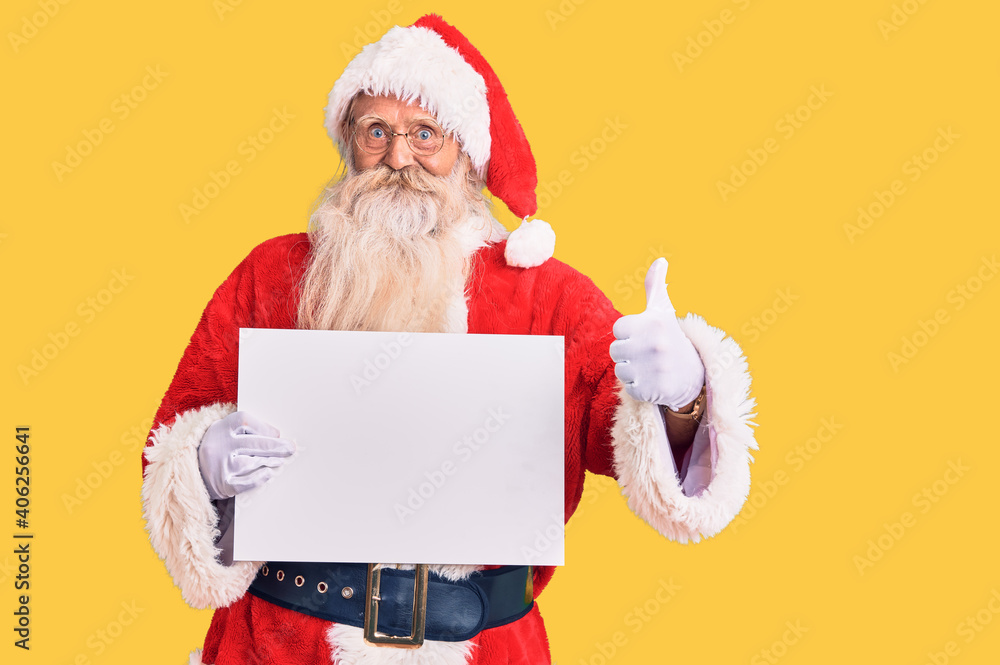 Old senior man with grey hair and long beard wearing santa claus costume holding banner smiling happy and positive, thumb up doing excellent and approval sign Old senior man with grey hair and long beard wearing santa claus costume holding banner smiling happy and positive, thumb up doing excellent and approval sign