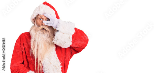 Old senior man with grey hair and long beard wearing traditional santa claus costume peeking in shock covering face and eyes with hand, looking through fingers with embarrassed expression.