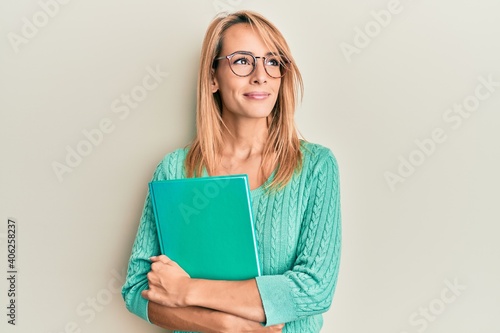Beautiful blonde woman holding book wearing glasses smiling looking to the side and staring away thinking.