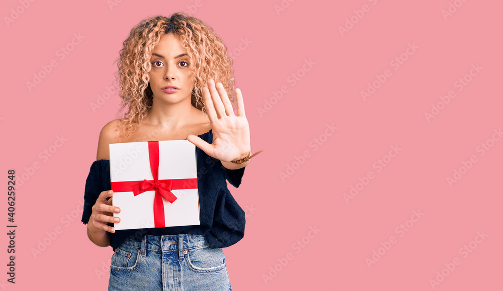 Young blonde woman with curly hair holding gift with open hand doing stop sign with serious and confident expression, defense gesture