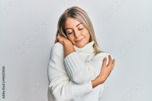 Beautiful blonde woman wearing casual turtleneck sweater hugging oneself happy and positive, smiling confident. self love and self care photo
