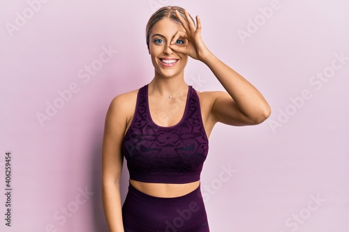 Beautiful blonde woman wearing sportswear over pink background doing ok gesture with hand smiling  eye looking through fingers with happy face.
