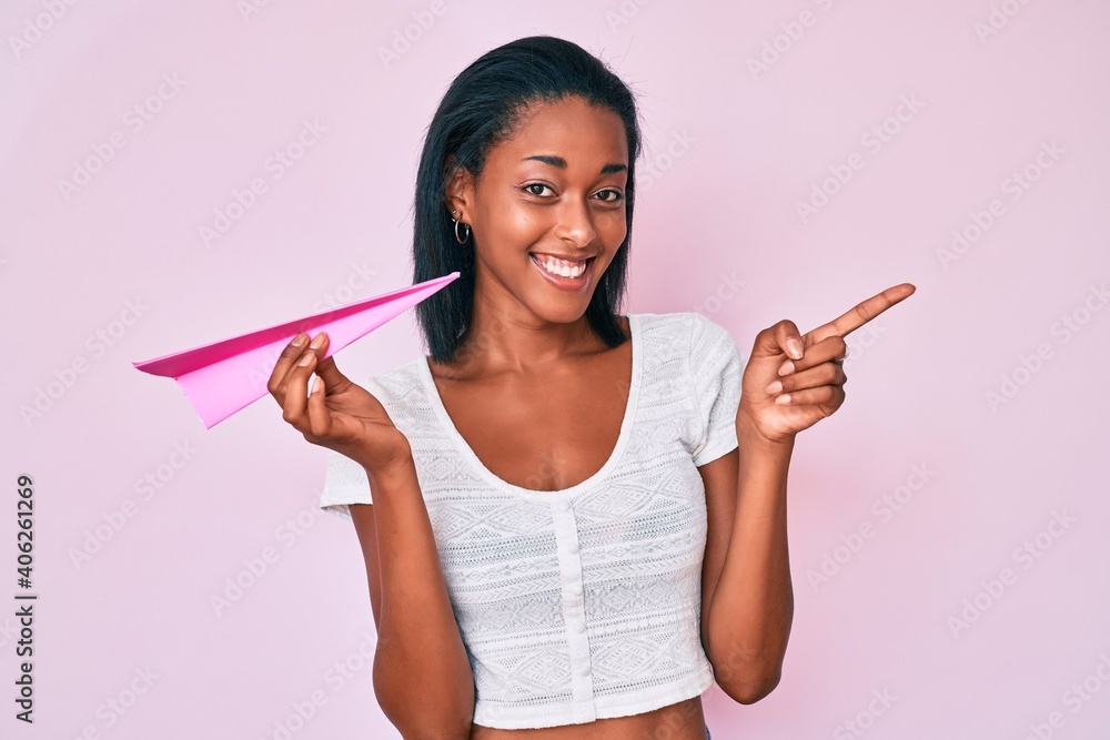 Young african american woman holding paper airplane smiling happy pointing with hand and finger to the side Young african american woman holding paper airplane smiling happy pointing with hand and finger to the side
