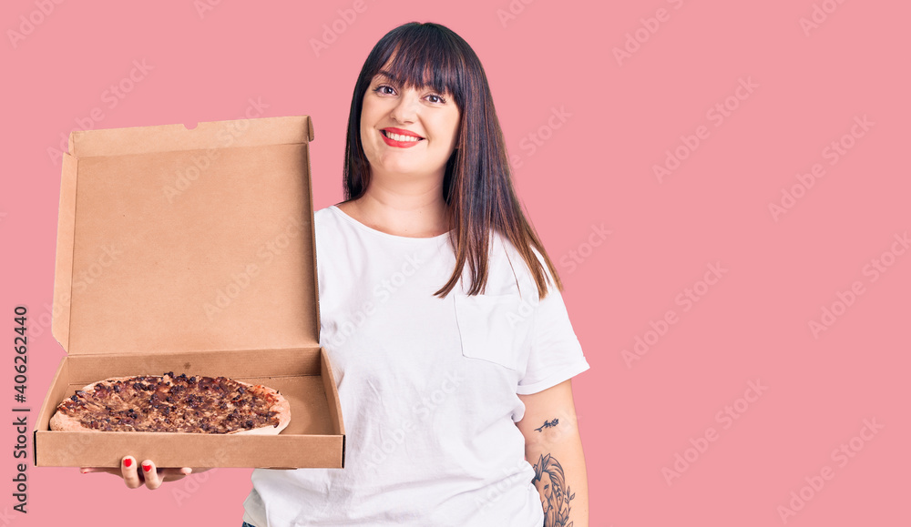 Young plus size woman holding delivery pizza box looking positive and happy standing and smiling with a confident smile showing teeth Young plus size woman holding delivery pizza box looking positive and happy standing and smiling with a confident smile showing teeth