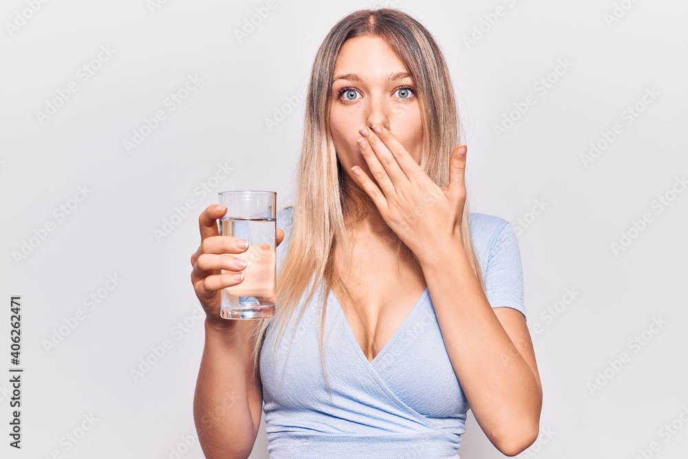 Young beautiful blonde girl drinking glass of water covering mouth with hand, shocked and afraid for mistake. surprised expression Young beautiful blonde girl drinking glass of water covering mouth with hand, shocked and afraid for mistake. surprised expression
