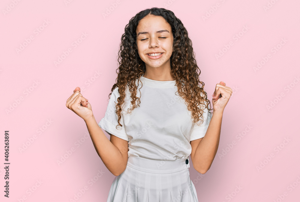 Teenager hispanic girl wearing casual white t shirt very happy and excited doing winner gesture with arms raised, smiling and screaming for success. celebration concept. Teenager hispanic girl wearing casual white t shirt very happy and excited doing winner gesture with arms raised, smiling and screaming for success. celebration concept.