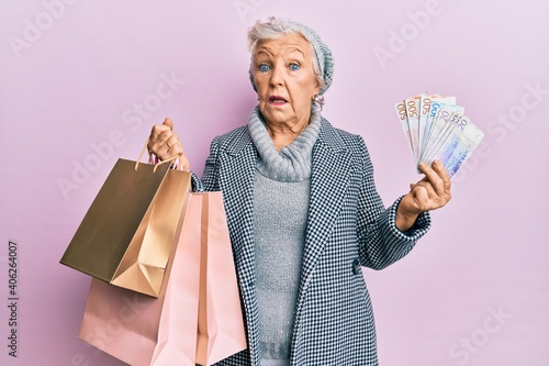 Senior grey-haired woman holding shopping bags and swedish krona banknotes clueless and confused expression. doubt concept.