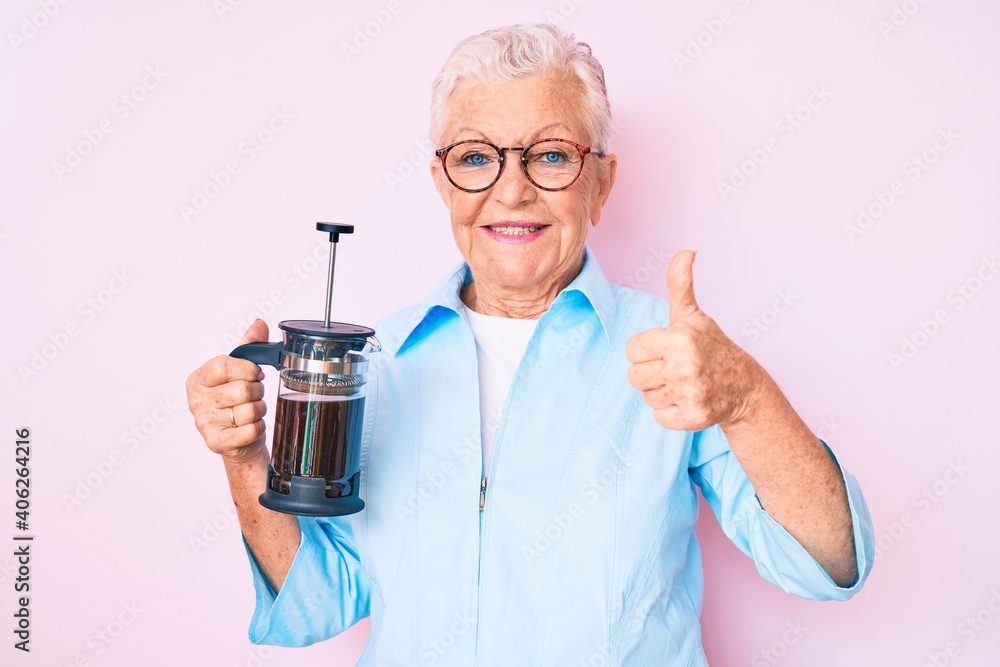 Senior beautiful woman with blue eyes and grey hair holding french coffee maker smiling happy and positive, thumb up doing excellent and approval sign Senior beautiful woman with blue eyes and grey hair holding french coffee maker smiling happy and positive, thumb up doing excellent and approval sign